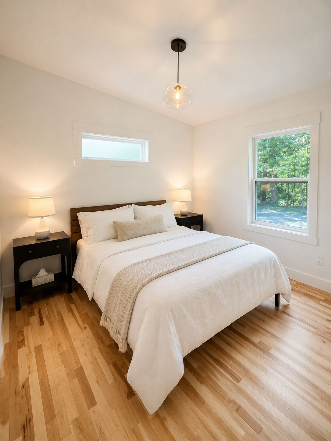 A bedroom with a wooden floor, a bed with white bedding, two bedside tables with lamps, a window with a view of greenery, and a ceiling light fixture.