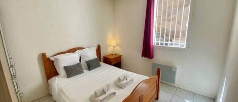 A bedroom with a wooden bed, white bedding, a nightstand with a lamp, a window with a view, and a radiator.
