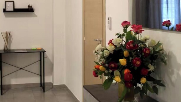 Een gang met een houten deur, een zwart metalen tafeltje met een bloemenvaas, een klein plankje met een fotolijstje en een grote spiegel aan de muur.