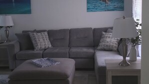 A living room with a grey sofa, a small ottoman, a white side table, and two wall-mounted lamps. - Coastal Retreat/ 2BR/2BA Split Floor Plan / Minutes to Anna Maria Island & Beach (Bradenton)