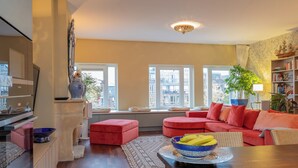 Ein Wohnzimmer mit einer Couch, Hocker, einem Esstisch mit Obstschale, einem Kamin und großen Fenstern.
