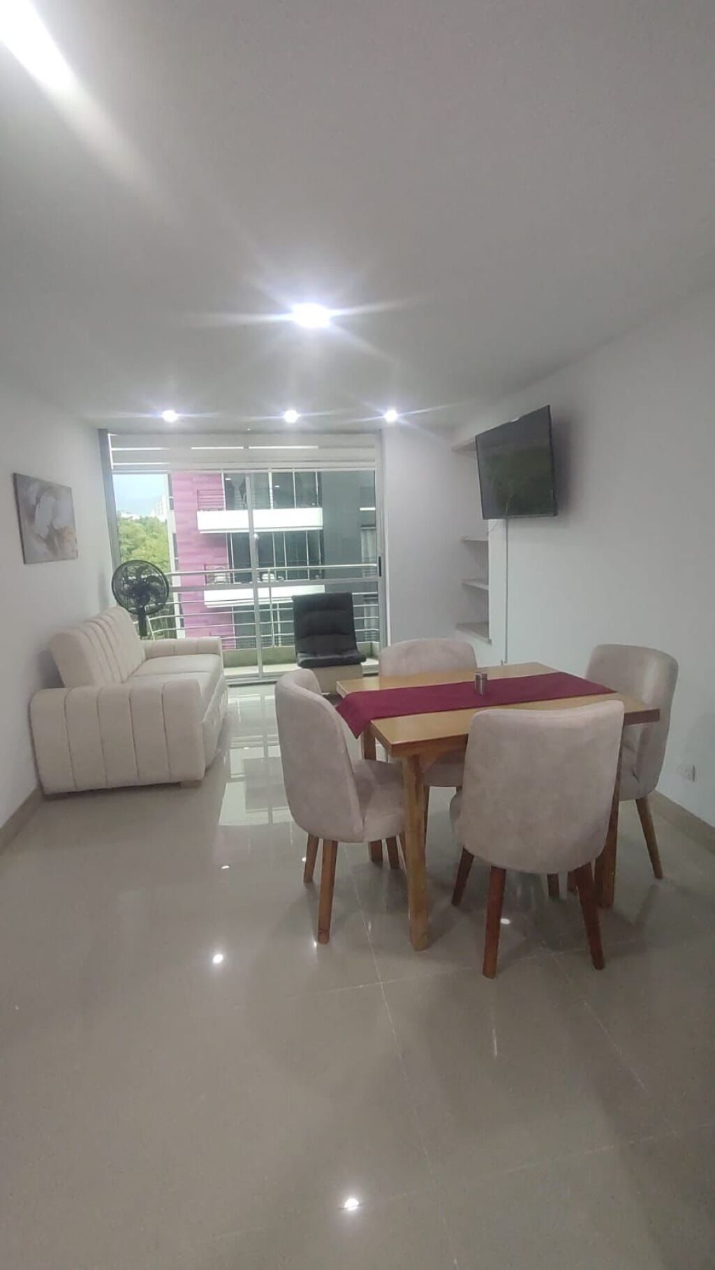 A modern living room with a dining area, a sofa, a TV mounted on the wall, and a balcony with a view of a building.
