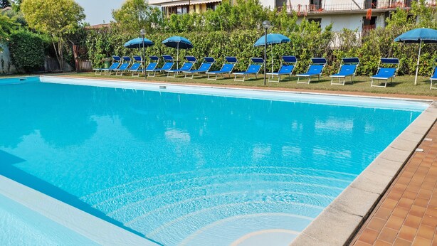 A swimming pool with blue lounge chairs and umbrellas, surrounded by greenery and buildings. - Apartment 'La Dolce Vacanza' with Private Terrace, Wi-Fi and Air Conditioning (Desenzano del Garda)