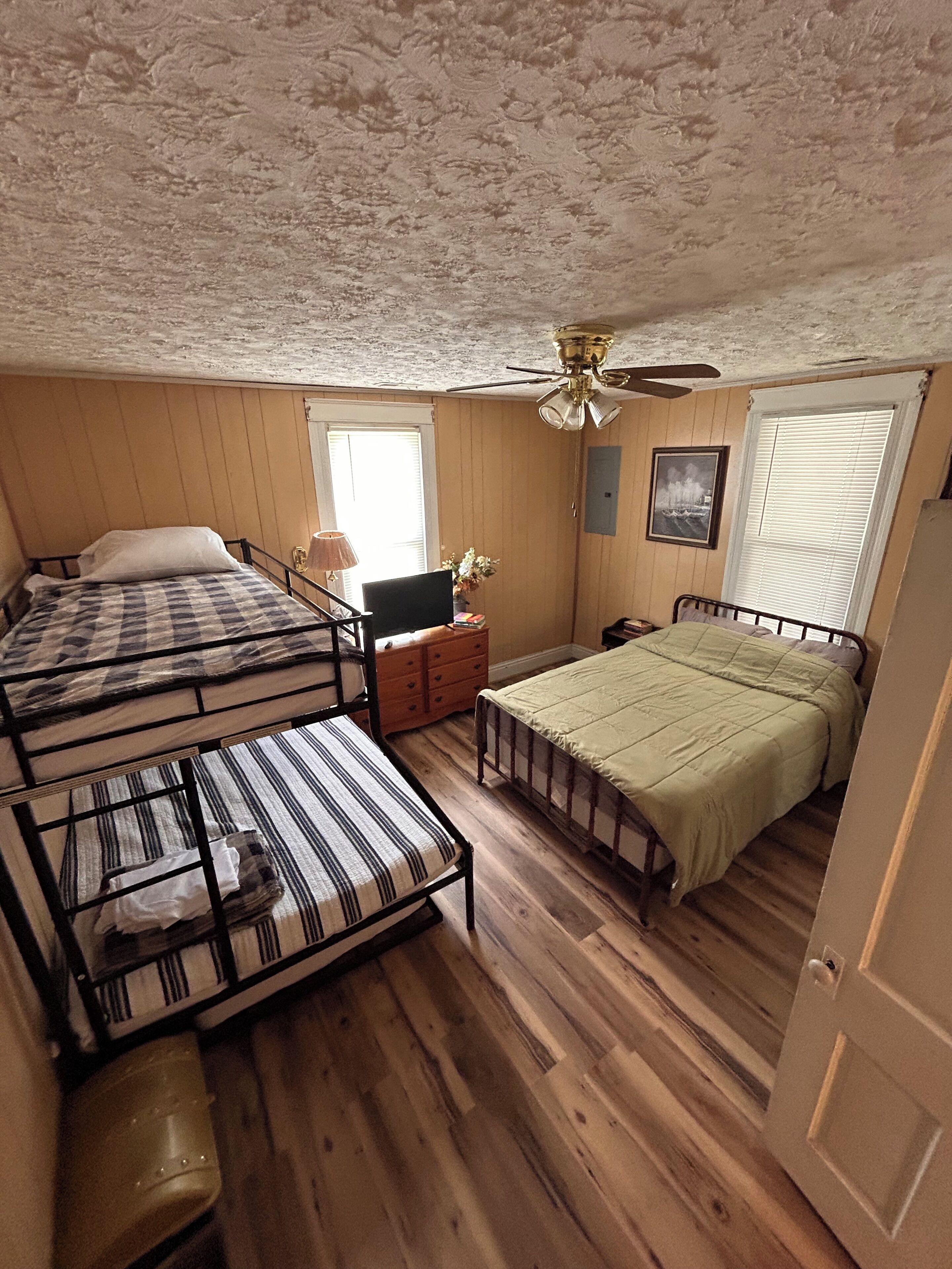 A room with two beds, a desk, a chair, a ceiling fan, and a window with blinds.