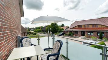Ein Balkon mit zwei Stühlen, einem Tisch und einem weißen Sonnenschirm, mit Blick auf eine Wohngegend mit Häusern und einem Auto.