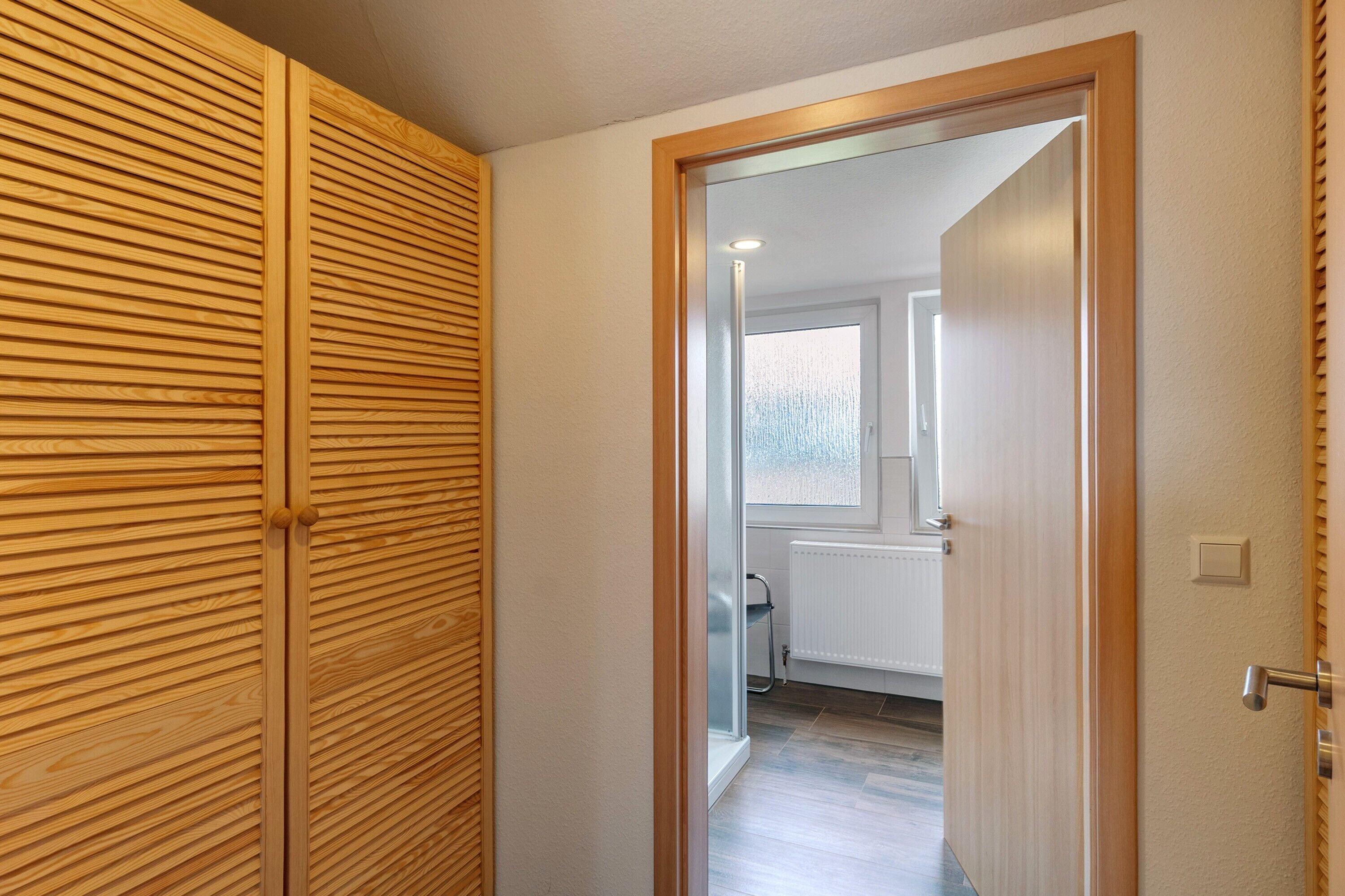 Ein Flur, der zu einem Badezimmer mit Heizkörper, Fenster und einer Tür mit Holzrahmen führt.