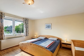 Ein Schlafzimmer mit einem hölzernen Bett, zwei Nachttischen mit Lampen, einem Fenster mit Vorhängen, einem Heizkörper und einem gerahmten Bild an der Wand.