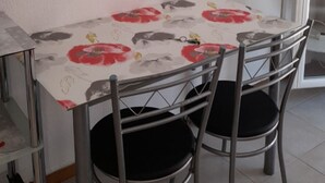 Ein Essbereich mit Tisch, der mit einer blumigen Tischdecke bedeckt ist, zwei Stühlen und einem an der Wand montierten Fernseher.