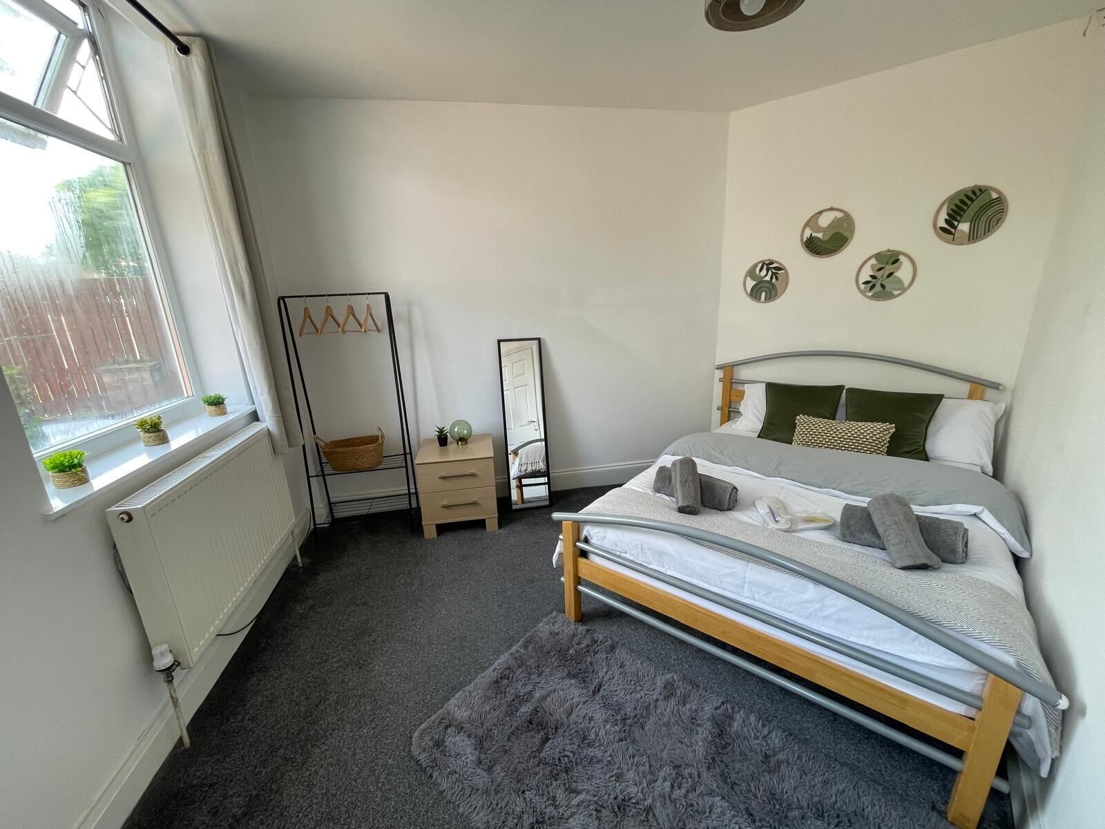A bedroom with a bed, a dresser, a mirror, and a window with plants.