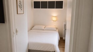 En smal hall leder in till ett sovrum med en säng som har en vit sänggavel, en sänglampa och ett element.