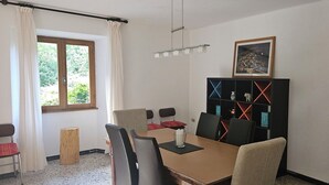 Ein Essbereich mit Tisch, Stühlen, einem Bücherregal, einem Fenster mit Vorhängen und einem gerahmten Bild an der Wand.