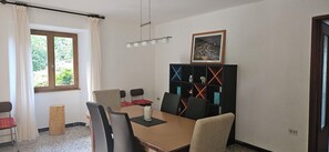 Uma sala de jantar com uma mesa, cadeiras, uma estante, uma janela com cortinas e um quadro na parede.
