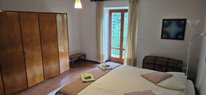 Quarto com cama, guarda-roupa de madeira, um banquinho, mesa de cabeceira e janela com cortinas.