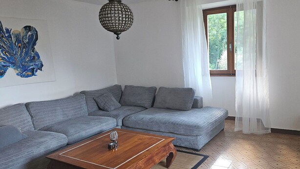 Ein Wohnzimmer mit grauem Sofa, einem Holztisch, einem Fernseher auf einem Schrank, einer Chandelieber und einem Fenster mit Vorhängen.