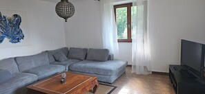 Un soggiorno con un divano grigio, un tavolino in legno, un televisore su un mobile, un lampadario e una finestra con tende.