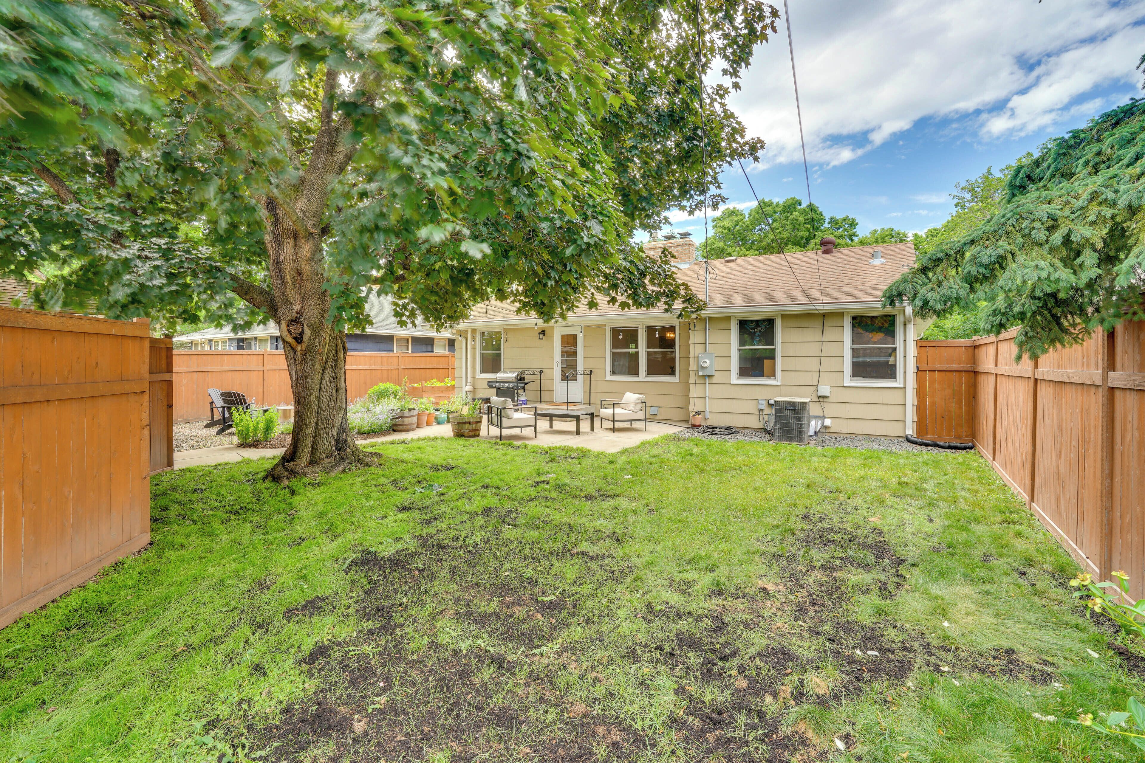 A backyard with a tree, a patio with outdoor furniture, and a wooden fence.