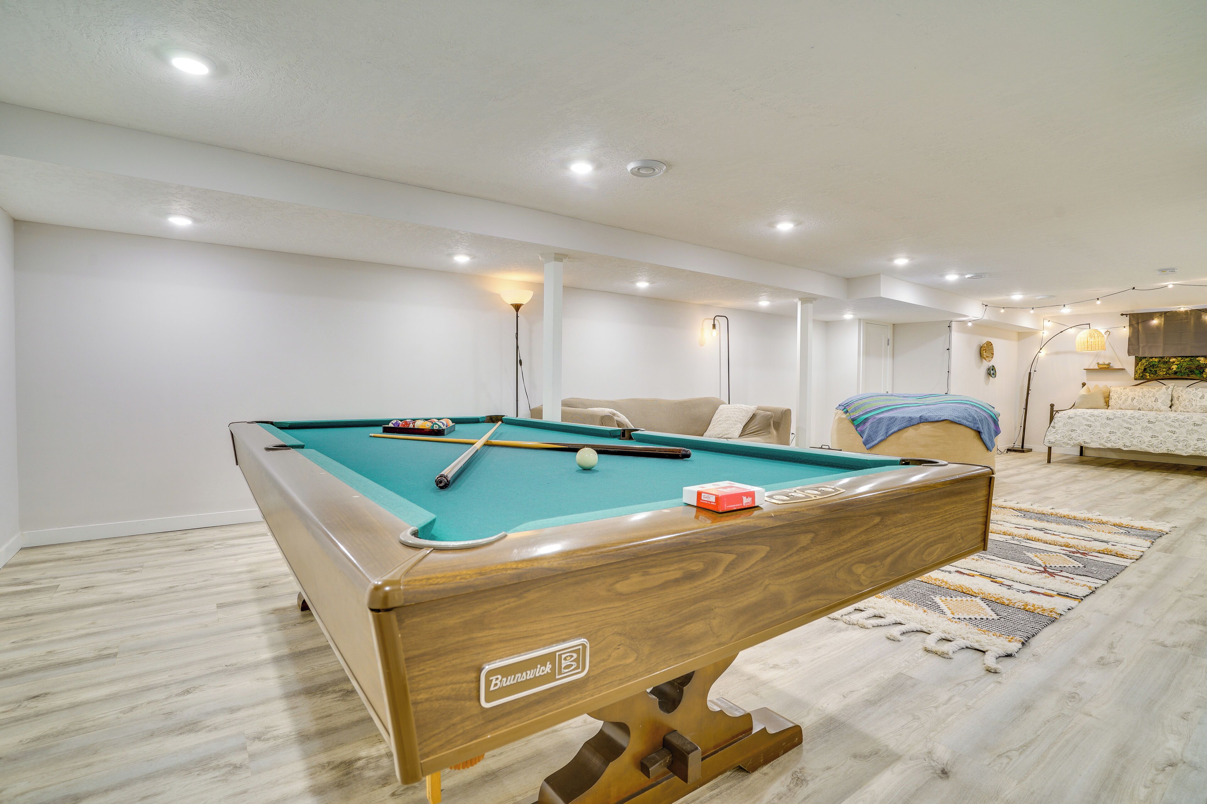 Una mesa de billar con bolas, un sofá y una cama en una habitación con paredes blancas y suelo de madera.
