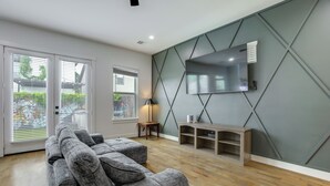 A modern living room with a sectional sofa, a flat-screen TV mounted on a geometric-patterned wall, and a wooden entertainment unit. - Game Room & Private Yard! Family Escape in Houston (Houston)