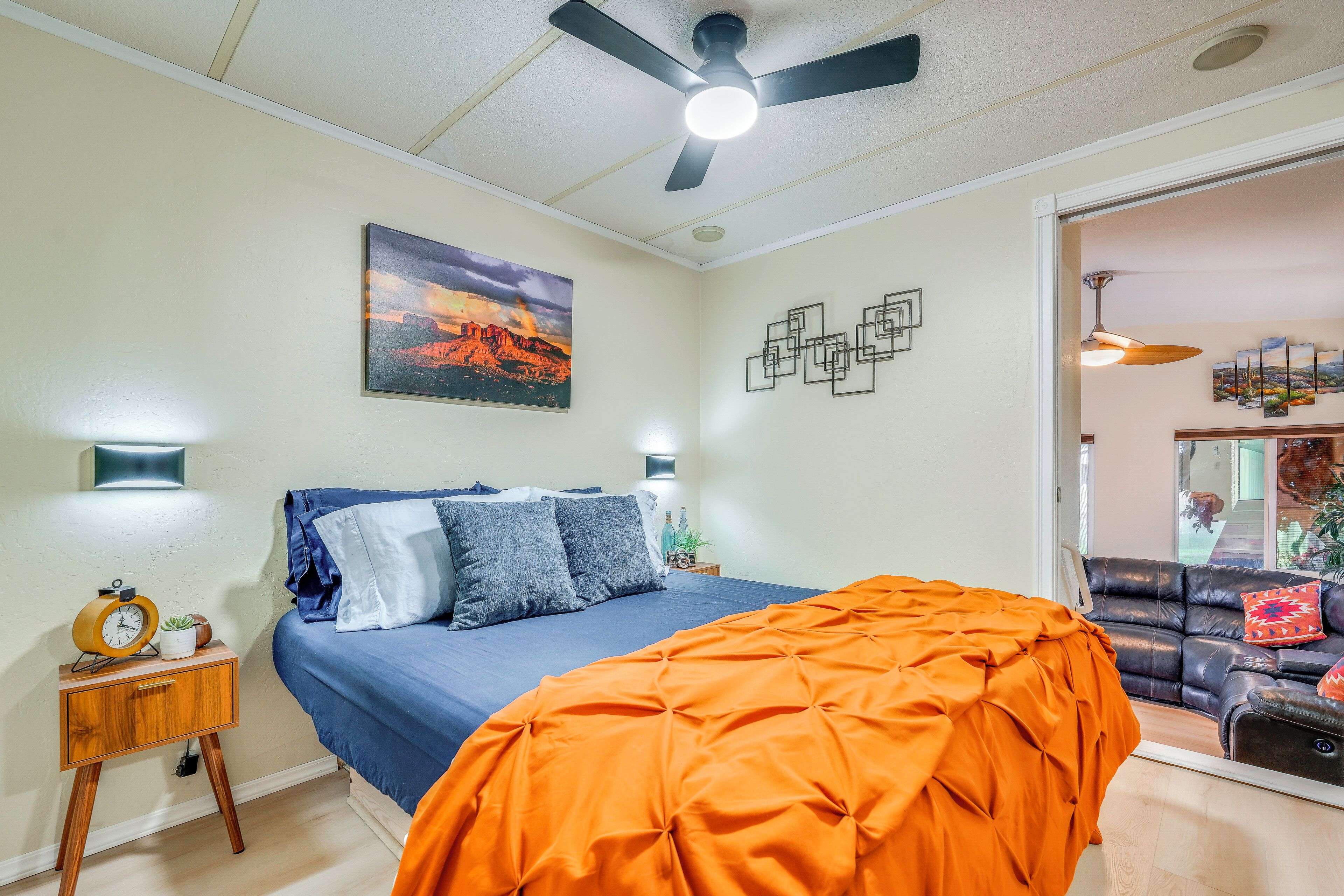 A bedroom with a bed, a bedside table, a ceiling fan, a wall-mounted lamp, and a painting.