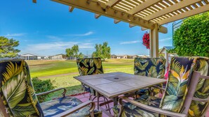 Une terrasse couverte avec une table et des chaises, une pergola au-dessus, et une vue sur une pelouse verdoyante et des maisons au loin.