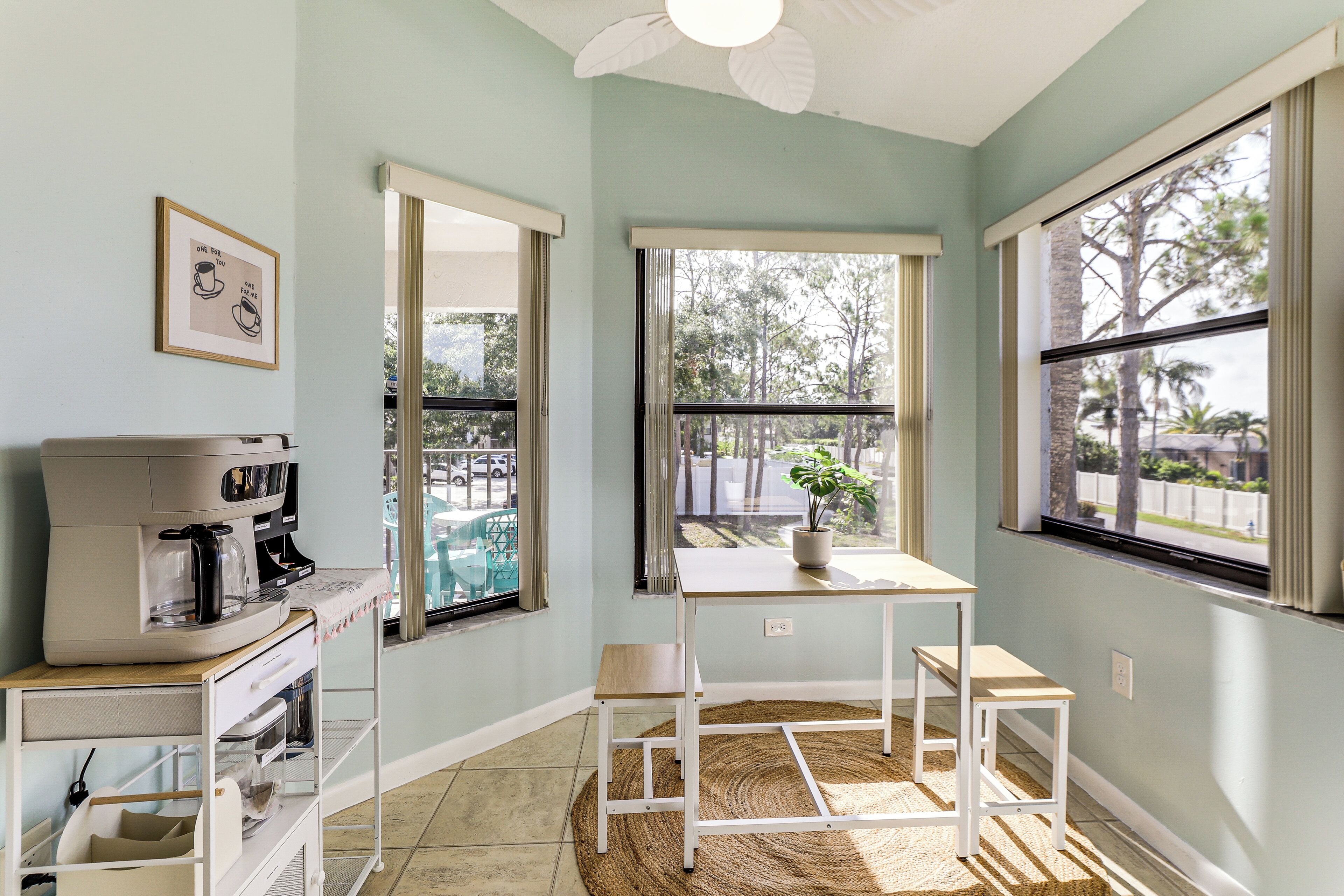 A kitchen with a coffee maker, a table, and chairs, a window with a view of trees, and a ceiling fan.