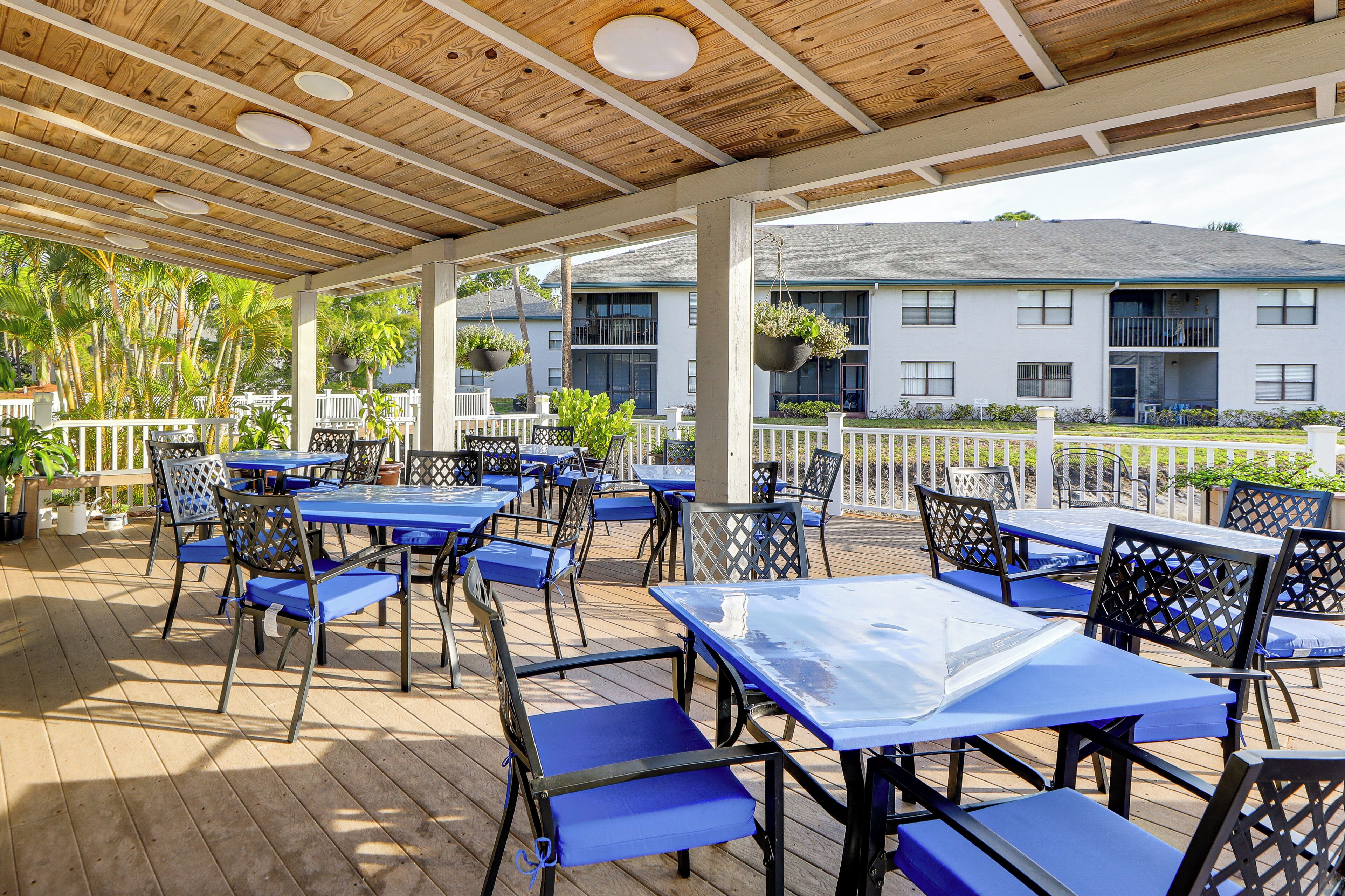 An outdoor patio area with tables and chairs, a wooden ceiling, and a view of a building in the background.