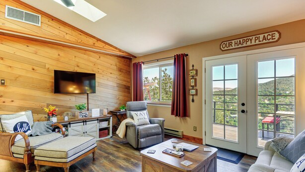 A cozy living room with a flat-screen TV, comfortable seating, a sign reading 'OUR HAPPY PLACE,' and a view through the door. - < 2 Mi to Whiskey Row! Mtn-view Adventure Basecamp (Prescott)