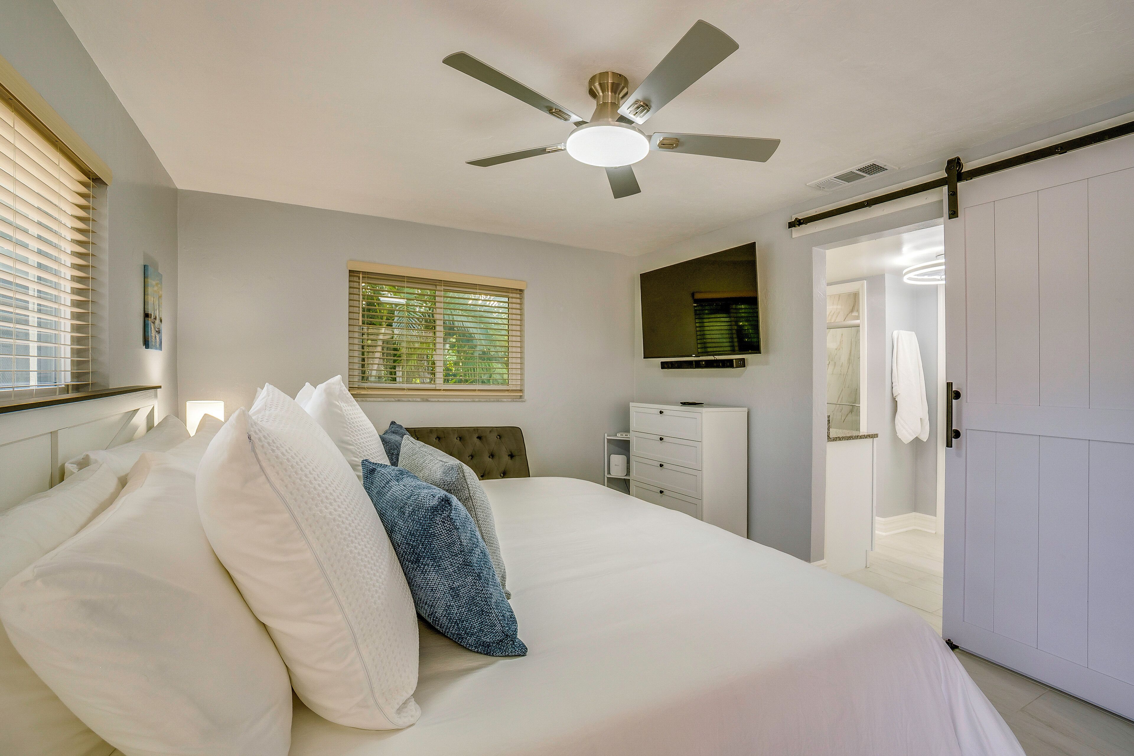 A bedroom with a bed, a ceiling fan, a window with blinds, a wardrobe, and a door leading to another room.