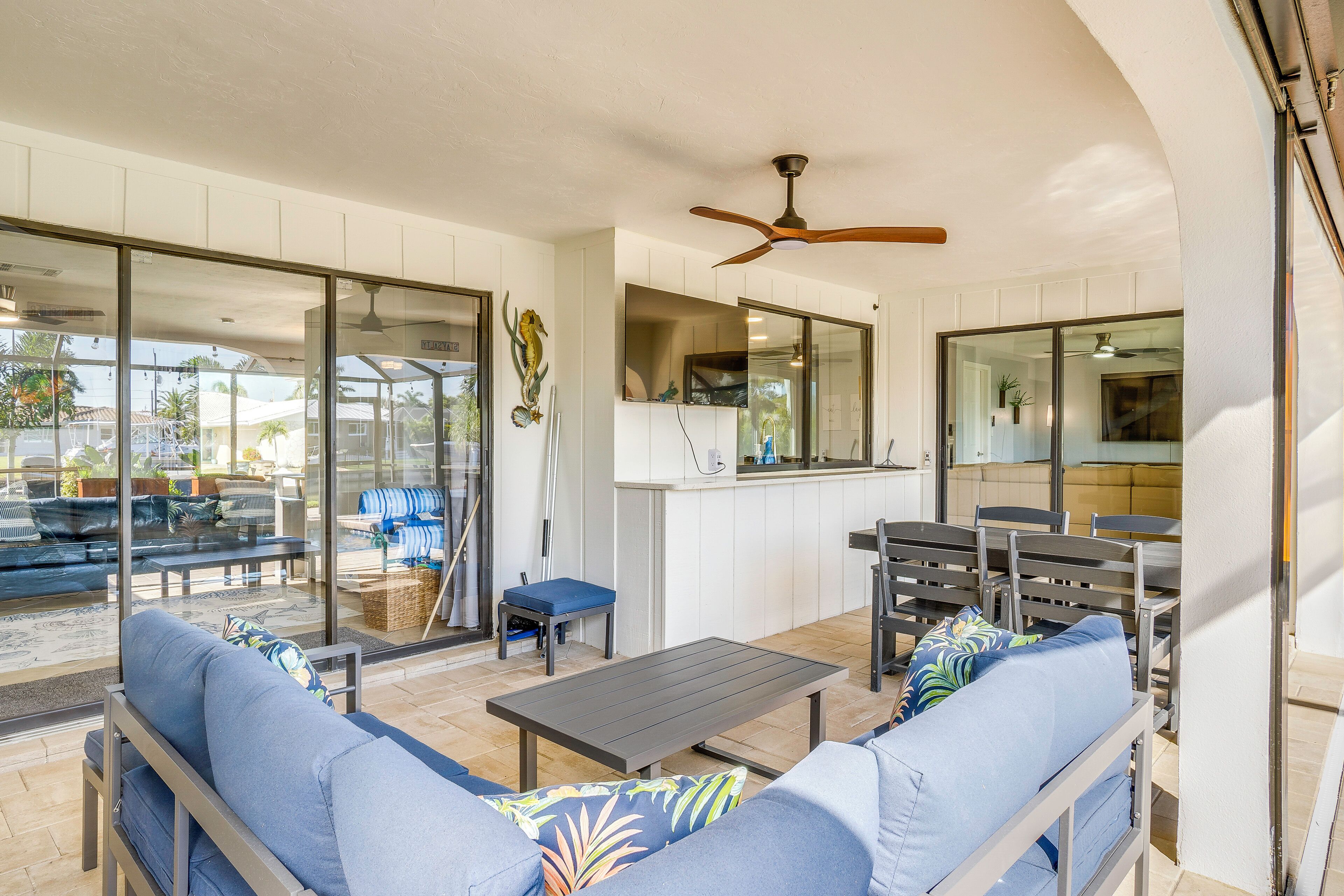 A modern outdoor living area with a sofa, table, and chairs, a ceiling fan, and a kitchen area visible through glass doors.