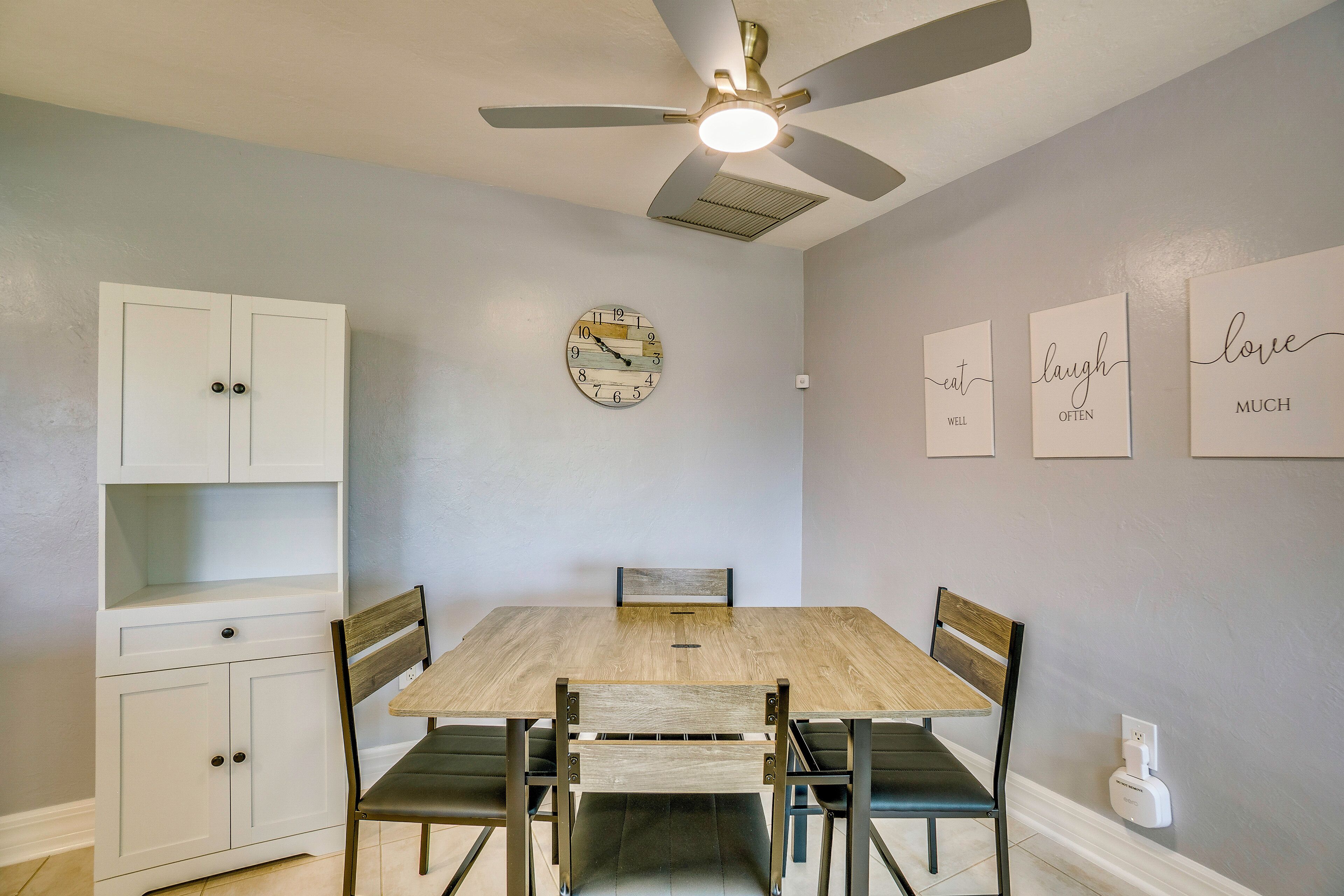A dining area with a wooden table, four chairs, a ceiling fan, a clock, and wall art.