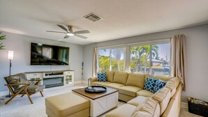 A living room with a sofa, ottoman, coffee table, TV, and a ceiling fan. - Waterfront Pgi, Heated Pool, Dock, 2 King Suites (Punta Gorda)