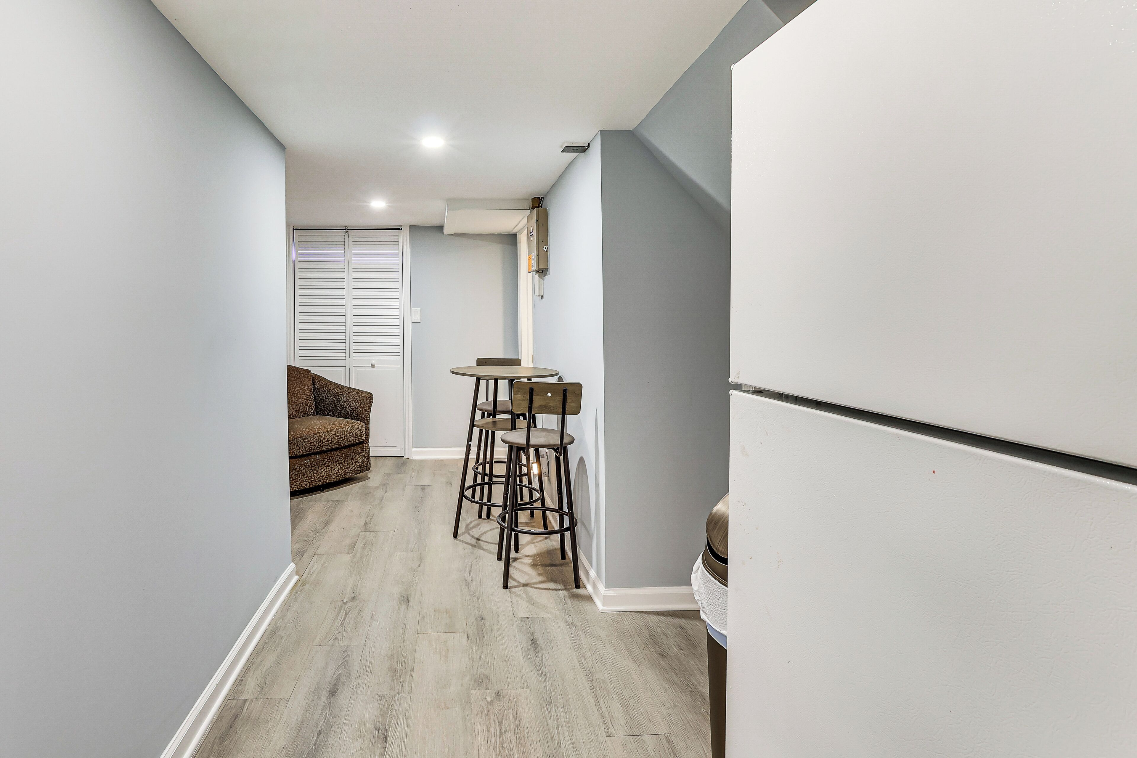A narrow hallway with a dining area featuring a table and chairs, a sofa, and a white refrigerator.