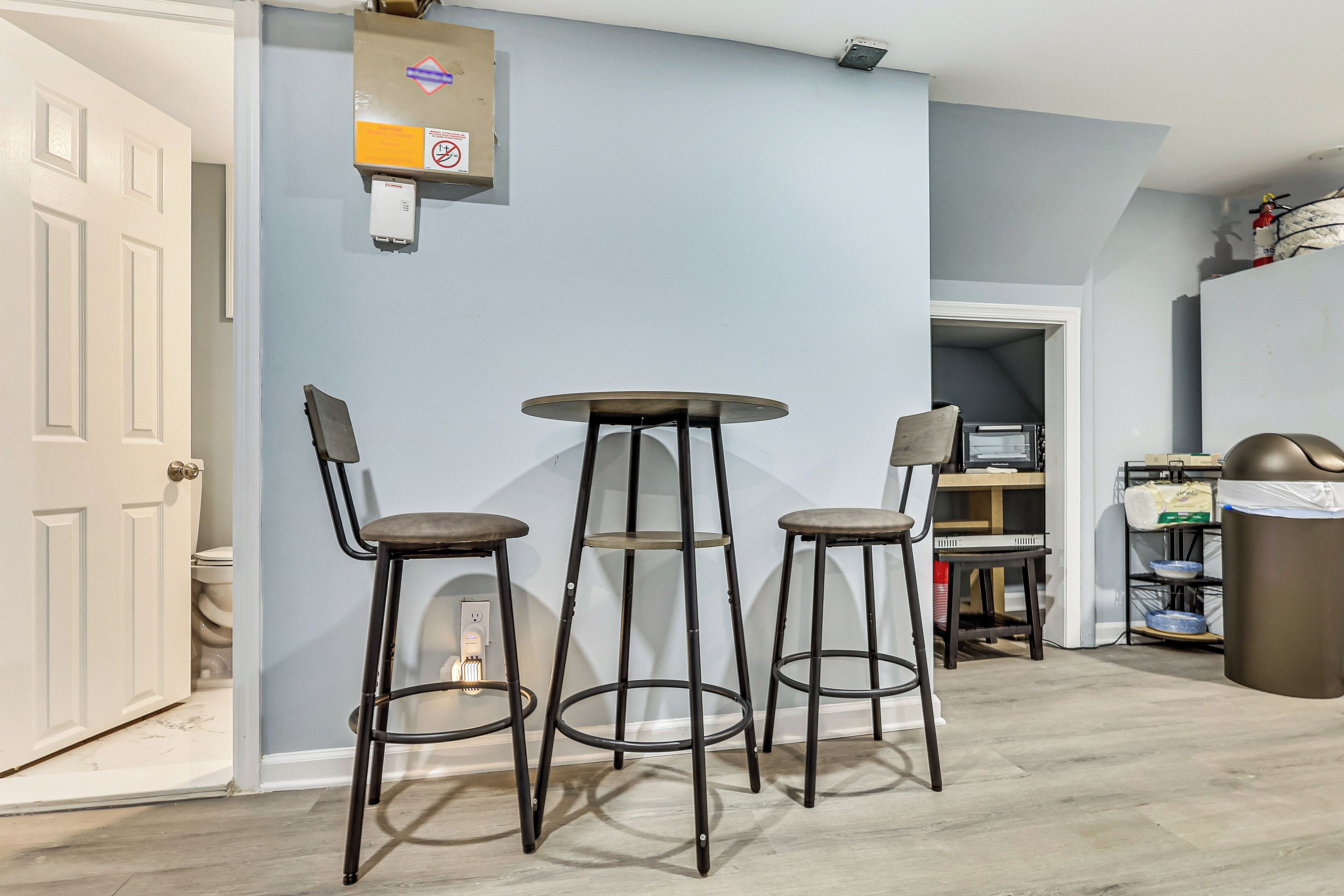 A room with a white door, a round table with two stools, a shelf with items, and a trash bin.