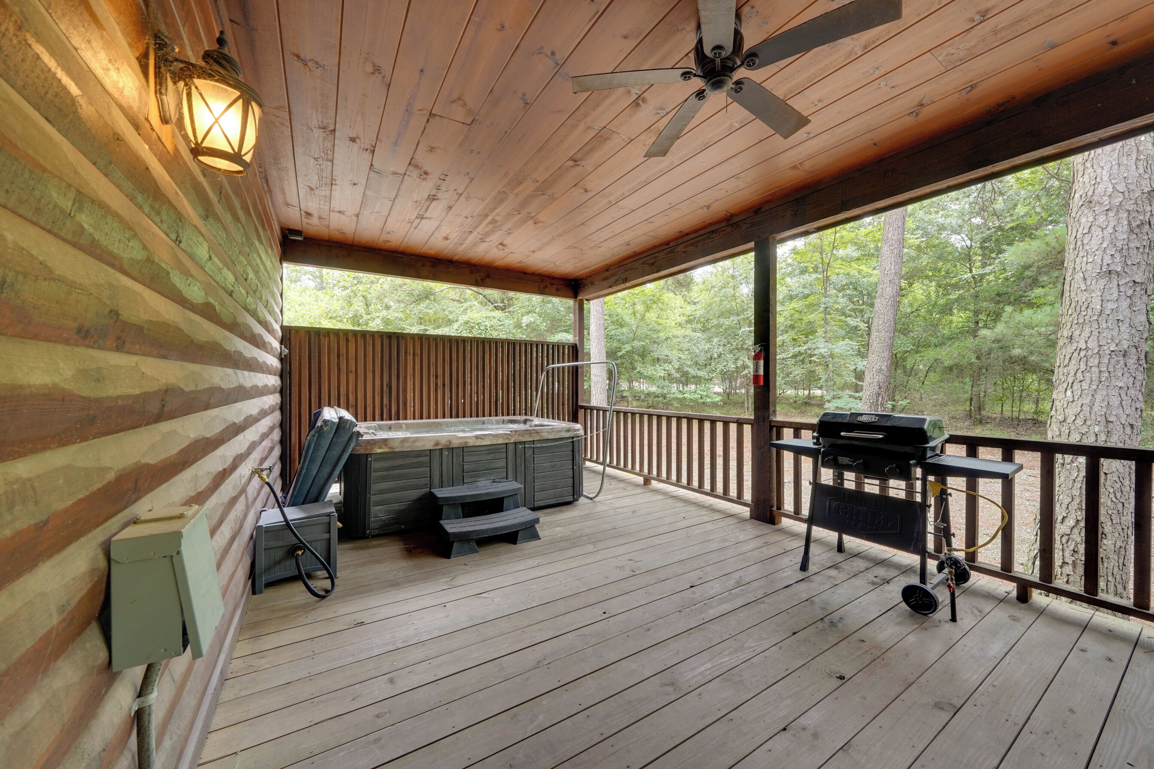 Une terrasse couverte avec un jacuzzi, un barbecue et un espace salon, entourée de murs en bois et d’arbres.