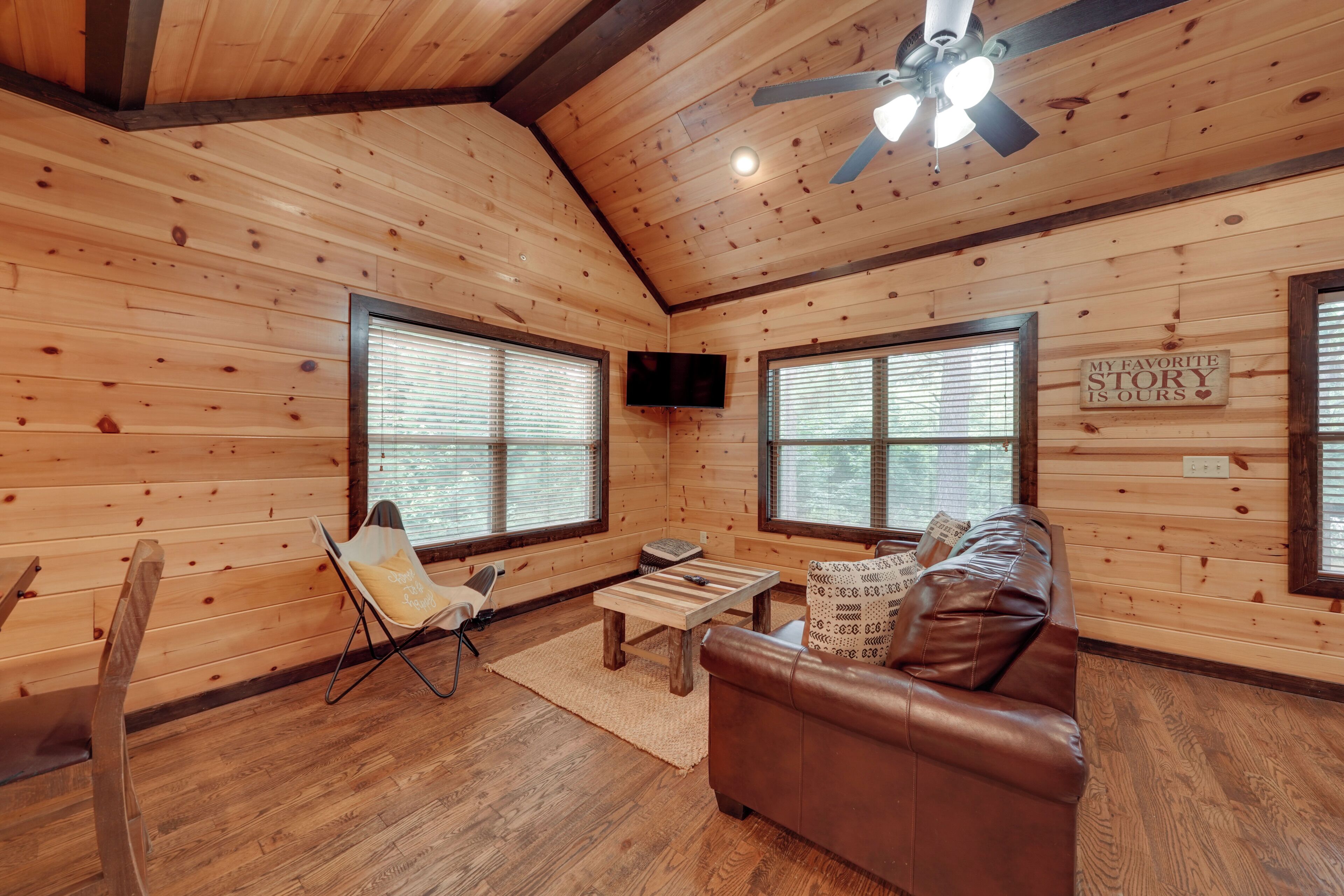 Interior acogedor de una cabaña con paredes de madera, un ventilador de techo, un televisor, un sofá de cuero marrón y una ventana con persianas.