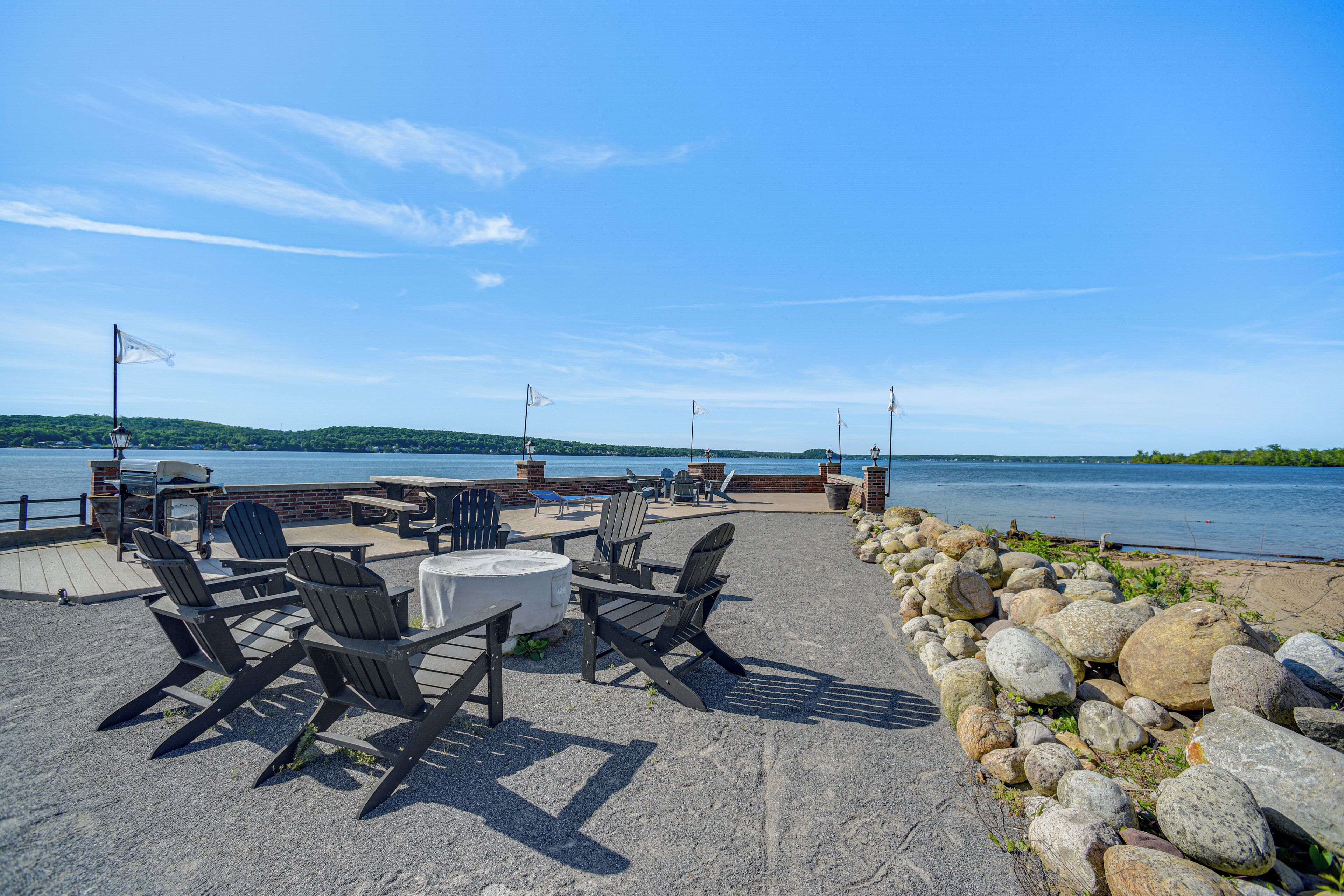 An outdoor seating area with Adirondack chairs and tables, a stone wall, and a view of the water.