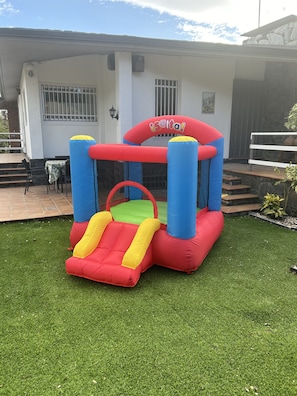 An inflatable bouncy house with a slide and a platform, set on artificial grass in front of a house with a stone facade and a staircase.