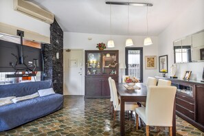 A modern living room with a sofa, dining table, chairs, and a kitchen area with a refrigerator, stove, and cabinets.