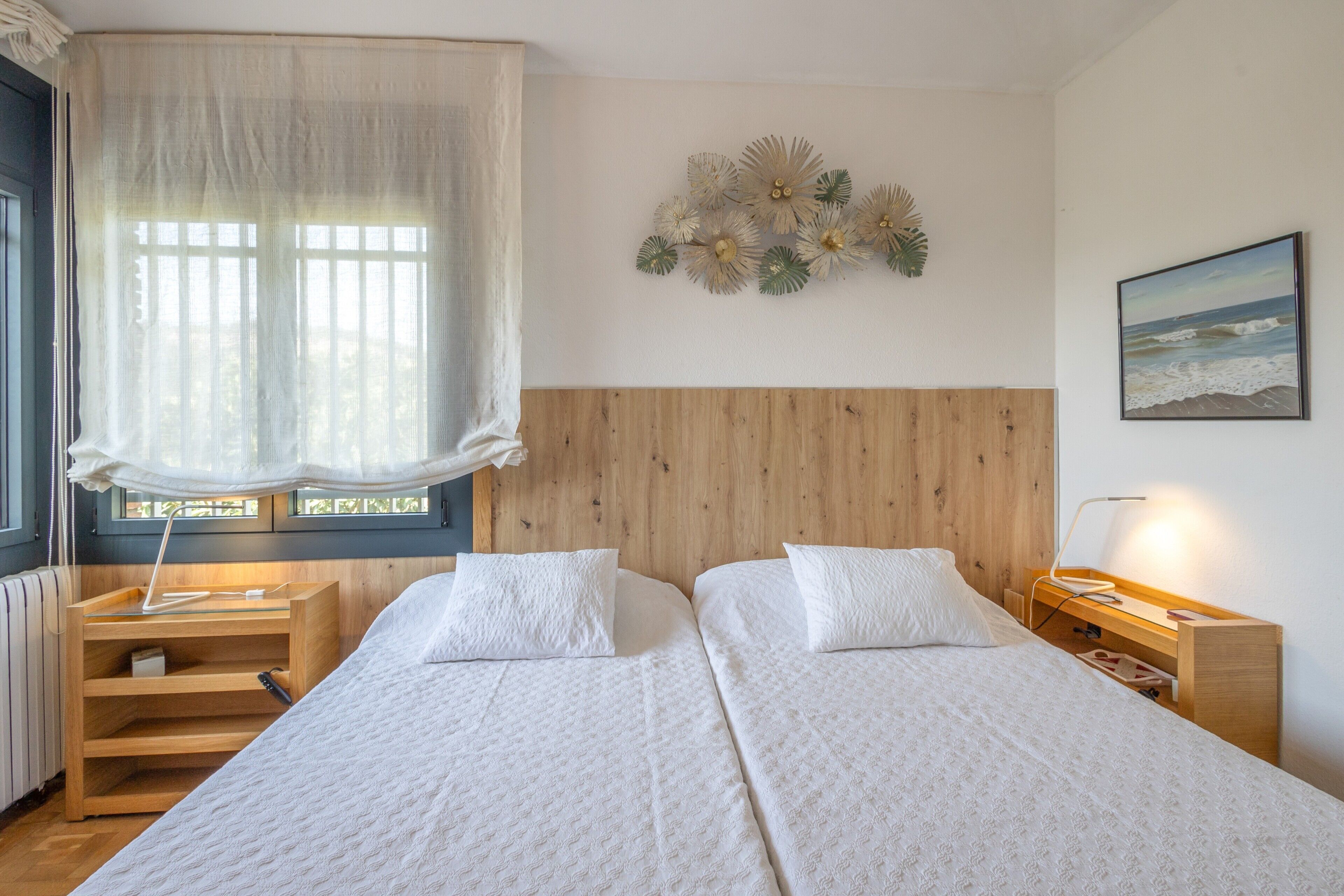Habitación de hotel con dos camas, cabecero de madera, ventana con cortinas translúcidas, mesita de noche con lámpara y un adorno en la pared.