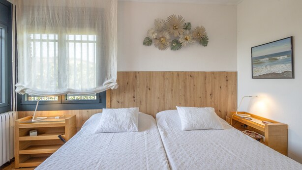 A hotel room with two beds, a wooden headboard, a window with sheer curtains, a bedside table with a lamp, and a wall-mounted decoration.