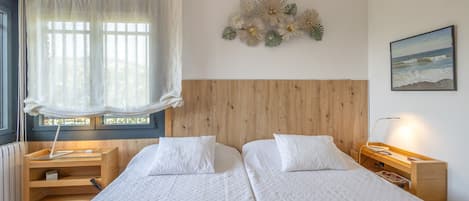 Habitación de hotel con dos camas, cabecero de madera, ventana con cortinas translúcidas, mesita de noche con lámpara y un adorno en la pared.