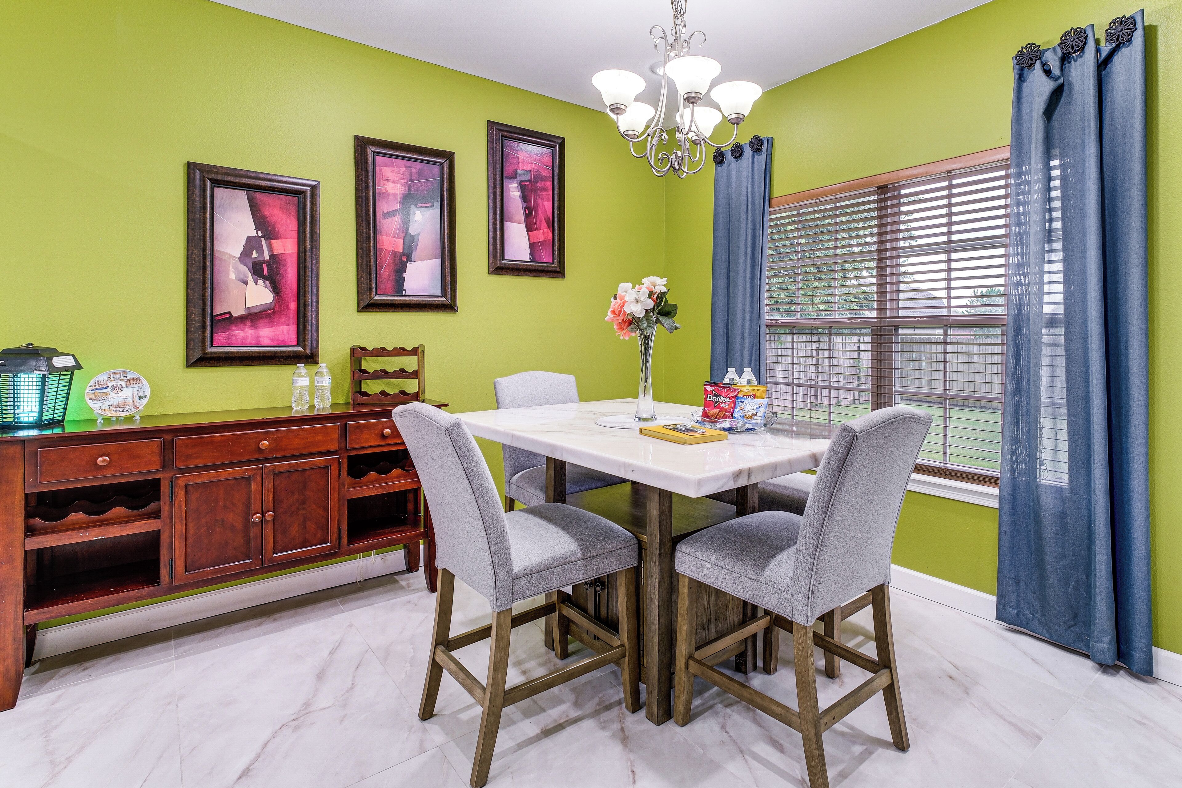 A dining area with a white table, grey chairs, a chandelier, and a window with blue curtains.