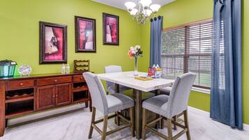 A dining area with a white table, grey chairs, a chandelier, and a window with blue curtains.