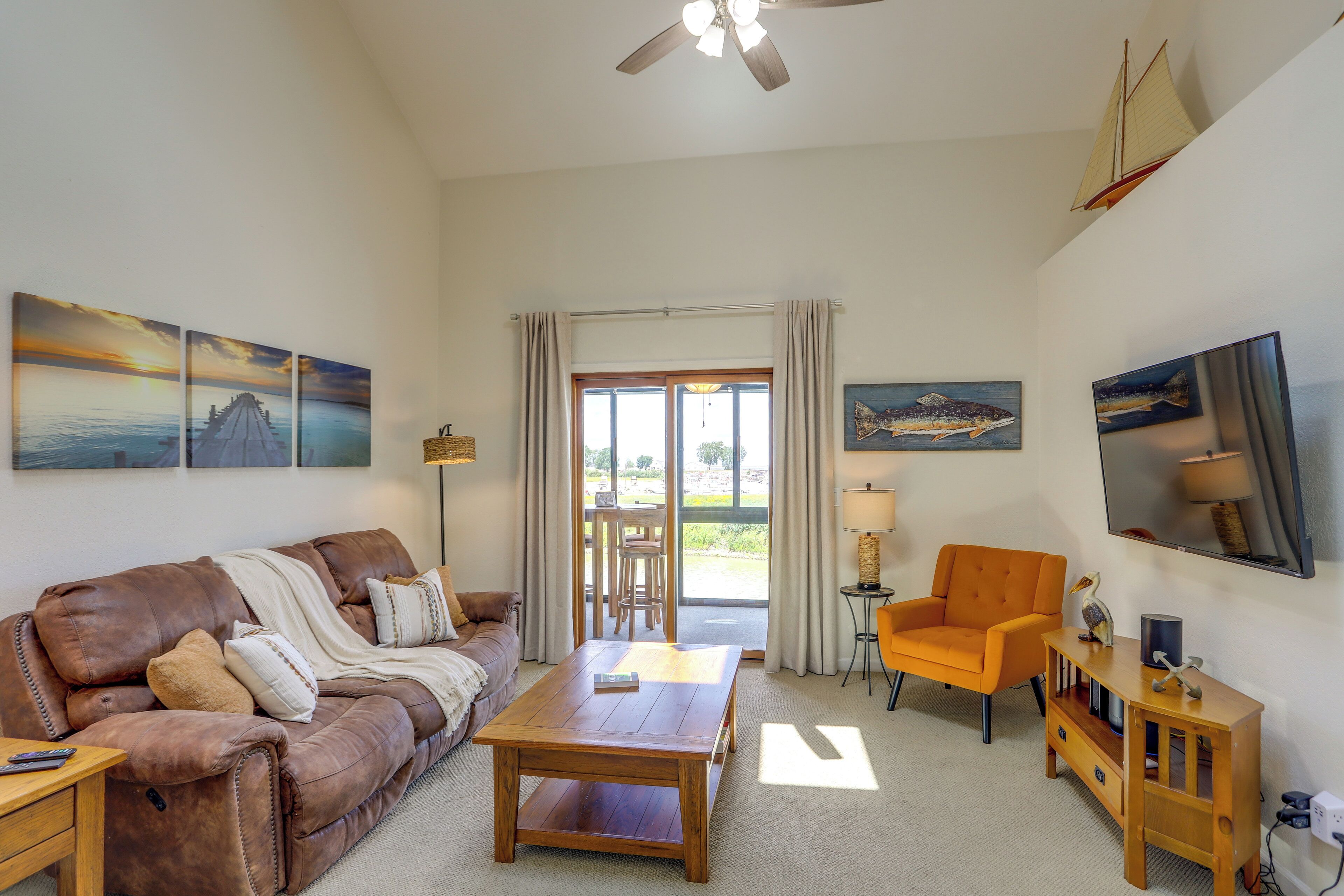A living room with a leather sofa, wooden coffee table, and an orange armchair. There is a large window with curtains, a ceiling fan, and artwork on the walls.