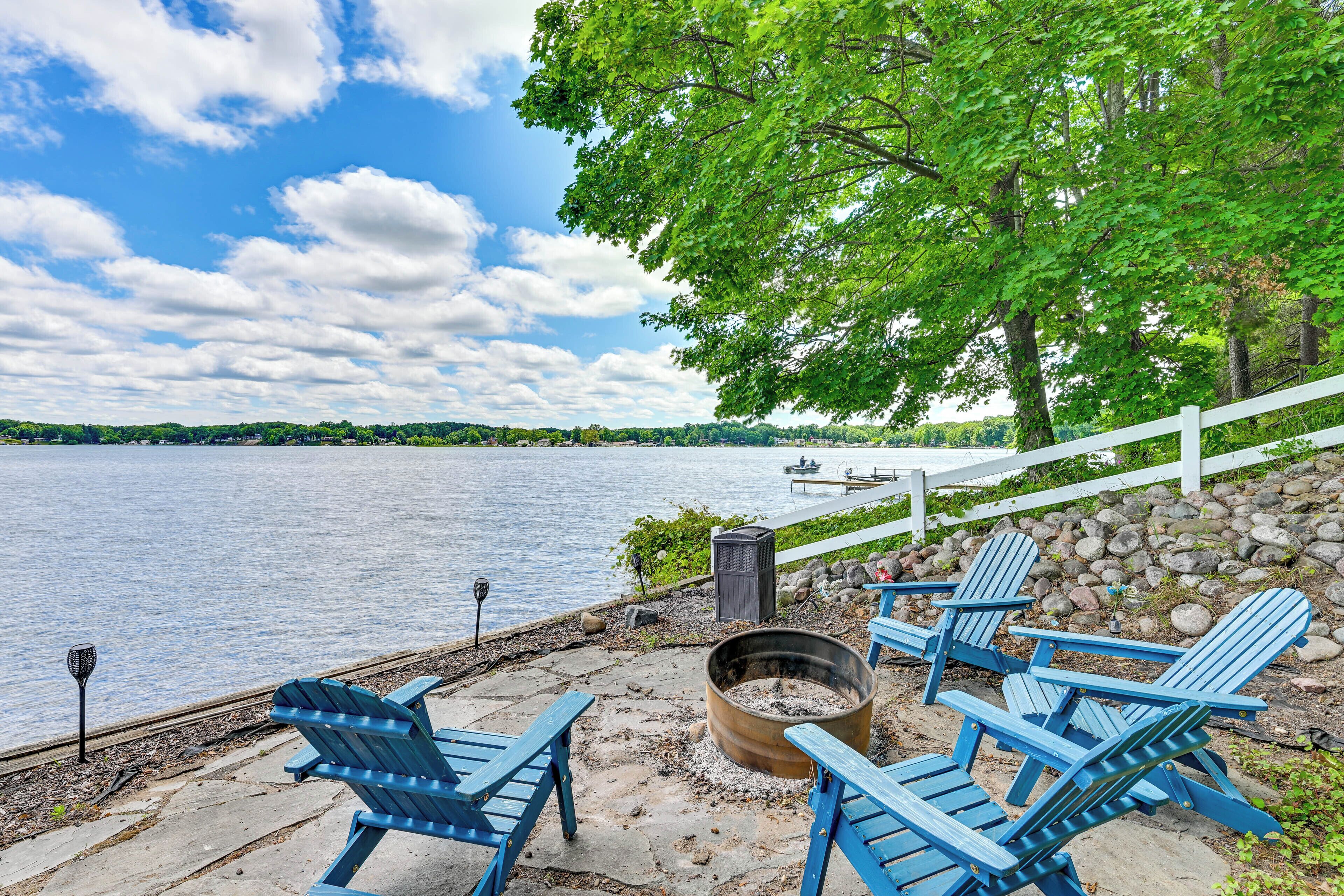 A lakeside deck with Adirondack chairs, a fire pit, and a white fence overlooking the water.