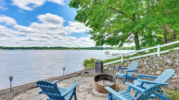A lakeside deck with Adirondack chairs, a fire pit, and a white fence overlooking the water.