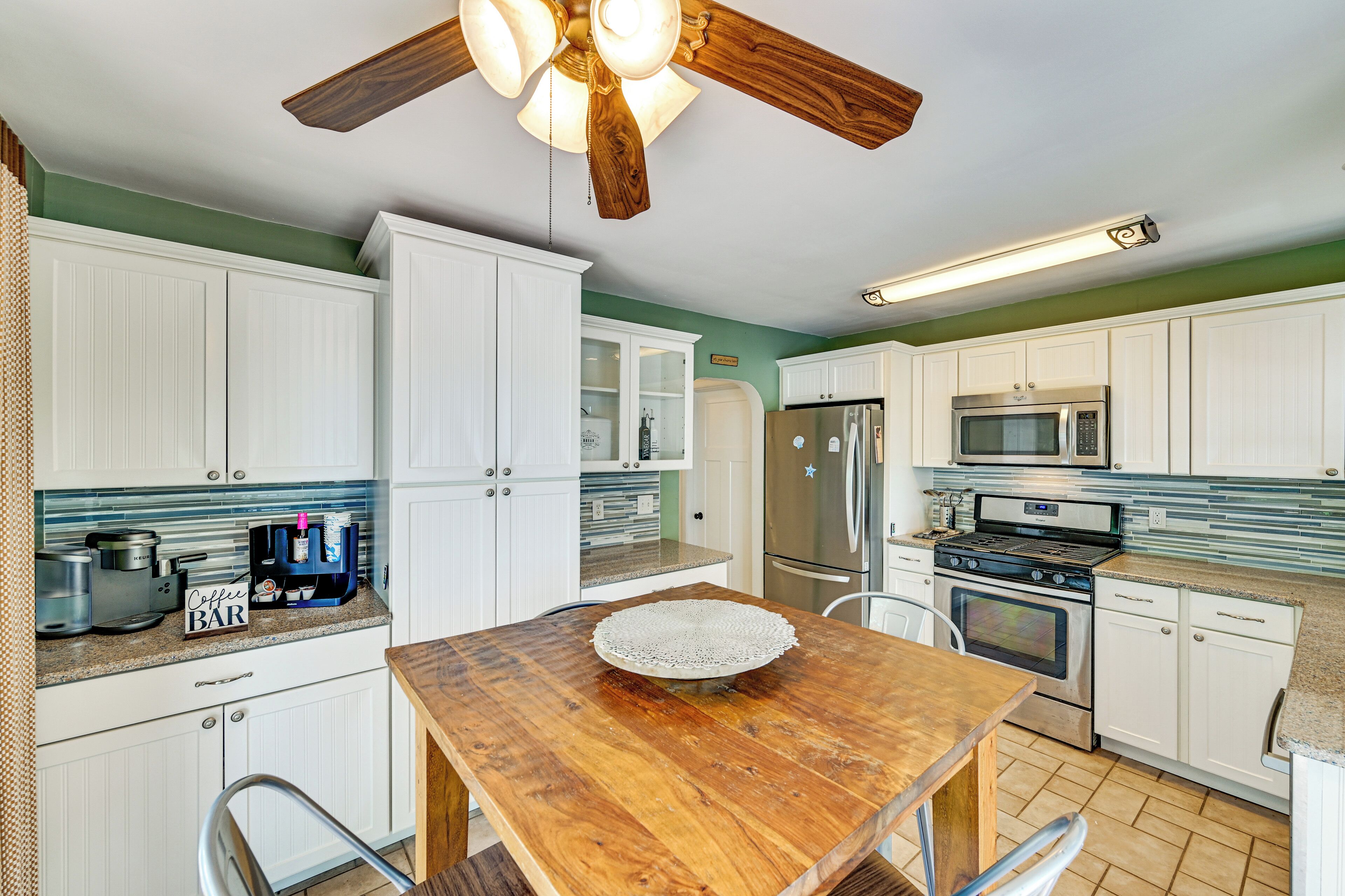A kitchen with white cabinets, a wooden table, a ceiling fan, and a coffee maker.