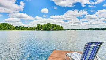 A wooden dock with a chair facing a lake, surrounded by greenery and trees.