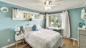 Un dormitorio con cama, mesita de noche, escritorio y silla, ventilador de techo y ventana con cortinas.