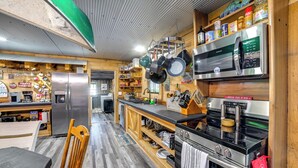 Ett kök med ett kylskåp, en mikrovågsugn och en spis i rostfritt stål, hyllor i trä med köksredskap och ett matbord i trä med stolar.